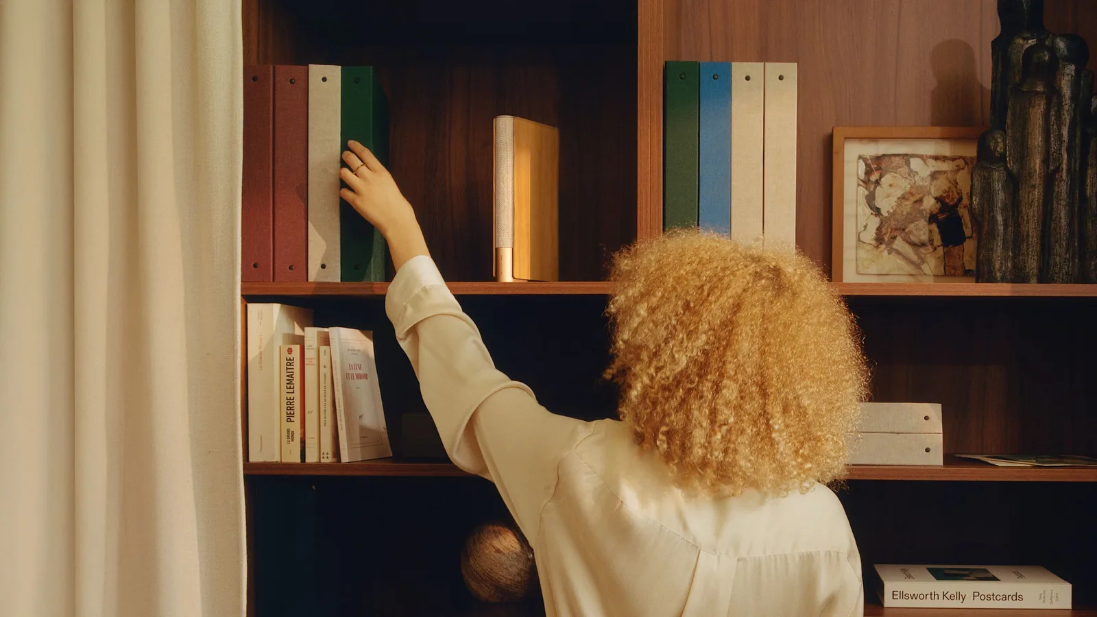 Bang & Olufsen Beosound Emerge Gold Tone speaker in gold-tone aluminium and oak, standing between books on a shelf